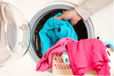 Mano mete ropa azul en lavadora. La ropa rosa y azul en la cesta sugieren que la lavadora huele mal.