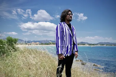 Uomo con giacca a righe blu e bianche guarda il mare in Sardegna, tra i campi di erba alta. Un'ambientazione perfetta per scoprire i **4 ristoranti Sardegna**.
