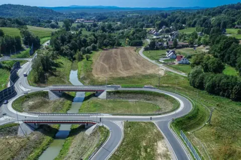 Nowoczesny węzeł drogowy na drodze krajowej 75, z mostami nad rzeką, otoczony zielonymi wzgórzami i domami.