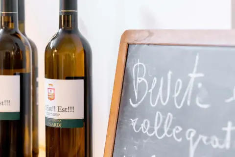Bottiglie di vino "Est!! Est!!!" in primo piano, con una lavagna che invita a "Bevete e rallegratevi".