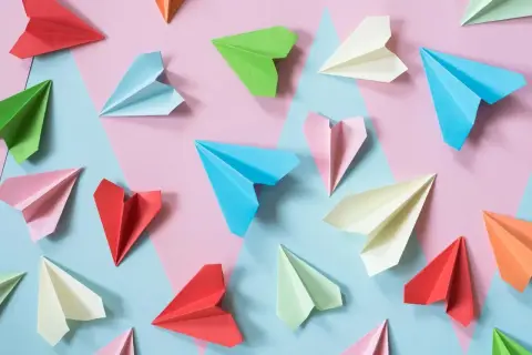 Kolorowe papierowe samolociki, jak się robi origami. Różne odcienie papieru ułożone na tle geometrycznych kształtów.