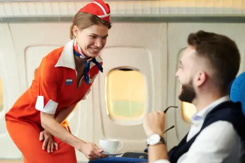 Uśmiechnięty stewardesa w pomarańczowym stroju podaje kawę pasażerowi w samolocie.