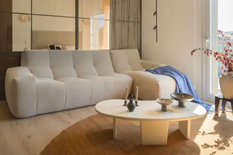 Divano chiaro e tavolino bianco su un tappeto di colore marrone. Sul tavolino, vasi e candele. Sul divano, una coperta blu.
