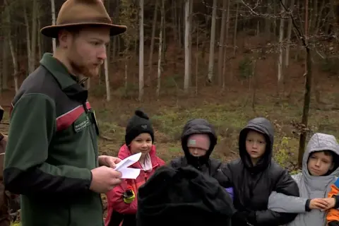 Ein Förster mit braunem Hut erklärt Kindern im Wald etwas. Sie sind von Hütten aus hierher gekommen, um mehr über die Natur zu lernen.