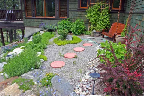 Inexpensive stepping stone walkway ideas: red circular pavers create a path through a gravel garden with lush greenery and a wooden chair.