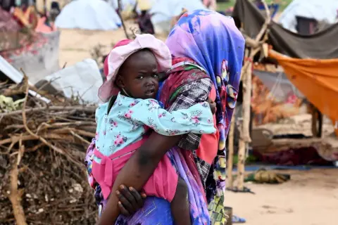 En kvinna bär ett barn i en flyktinglägerliknande miljö. Detta ger en inblick i fakta om Sudan och dess utmaningar.