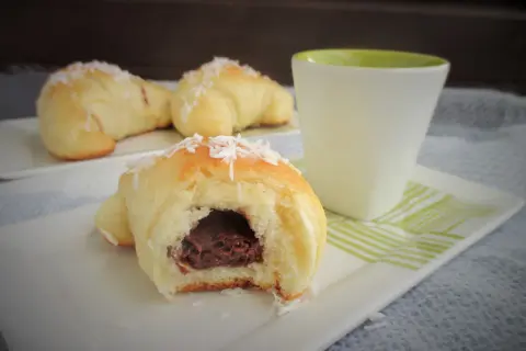Pyszne rogaliki drożdżowe z budyniem, posypane wiórkami kokosowymi, obok filiżanki.
