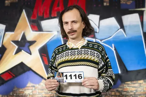 Mężczyzna z wąsami i w swetrze z geometrycznym wzorem trzyma tabliczkę z numerem 1116. Savyan czeka na swój występ.