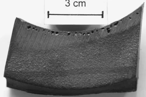 Fragment metalu z licznymi porami, widoczne wady odlewnicze. Skala 3 cm.