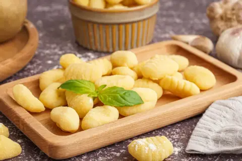 Domowe gnocchi, idealne do przygotowania według prostego przepisu. Na drewnianej desce świeże kluseczki ziemniaczane, ozdobione listkiem bazylii.
