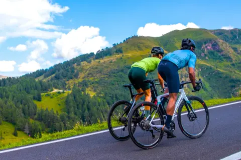 Dwóch kolarzy w sportowych strojach pokonuje górską trasę. Ich pozycja na rowerze szosowym jest aerodynamiczna, gotowi na wyzwanie.