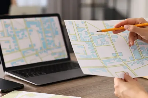 Mano con matita analizza mappa con edifici, simile a una **categoria catastale B4**, accanto a un laptop con mappa digitale.