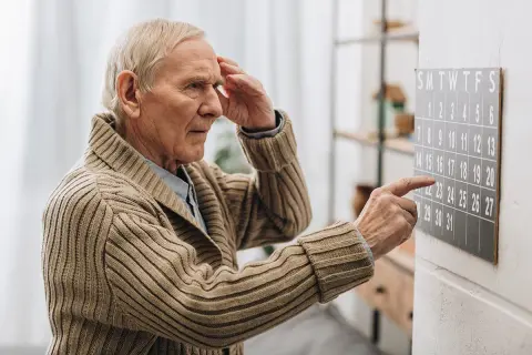 Älterer Mann mit Hand an der Stirn blickt auf Kalender. Ein Zeichen für Vergesslichkeit im Alter, wenn Daten verschwimmen.