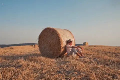 Jonge man met hoed leunt tegen hooibaal in een veld, lijkt verslaafd aan prikkels door de rust.