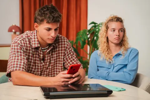 Een jongeman kijkt geconcentreerd naar zijn telefoon, terwijl zijn partner met gekruiste armen en een norse blik wegkijkt. Het voelt alsof hij haar compleet negeert, alsof ze leeggezogen wordt door zijn onoplettendheid.