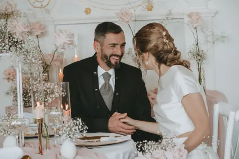 Ein glückliches Brautpaar bei ihrer Petersilienhochzeit, umgeben von zarten rosa Rosen und Schleierkraut. Sie halten Händchen und blicken sich liebevoll an.
