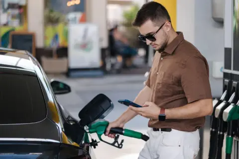 Un homme fait le plein de sa voiture à une **téléphone station essence**, consultant son smartphone.