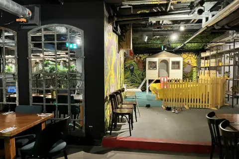 Warszawska restauracja z salą zabaw dla dzieci. Widać stoliki, krzesła i kolorowy plac zabaw z domkiem i zjeżdżalnią.