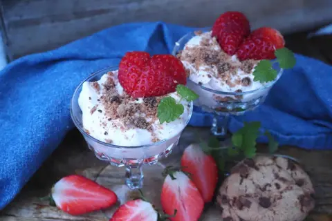 Två glas med krämig dessert toppad med jordgubbar och smulor. Bredvid ligger halverade jordgubbar och chokladkakor. En härlig sommardessert med jordgubbar och glass.