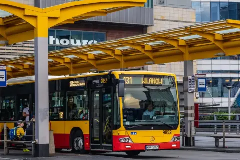 Żółto-czerwony autobus miejski na przystanku. Kierowca autobusu, jak i pasażerowie, zastanawiają się, ile zarabia kierowca autobusu.