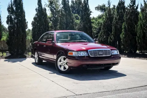 Bordowy Ford Victoria z chromowanym grillem stoi na betonowym podjeździe, otoczony przez wysokie, ciemnozielone drzewa.