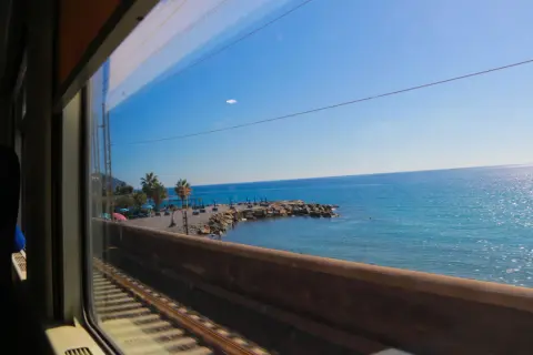 Vy från tågfönstret under en resa för att tågluffa i Italien. Blå himmel, glittrande hav och en stenstrand med palmer.