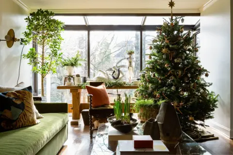 Cozy apartment Christmas decor features a decorated tree, green sofa, and a large window overlooking trees.