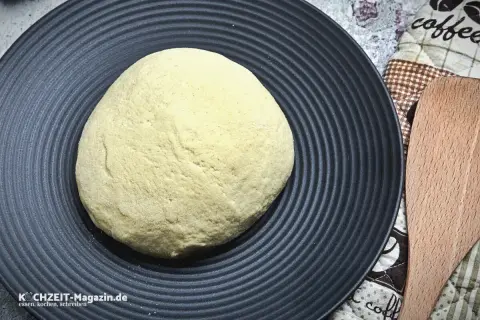 Ein runder, heller pizzateig wie beim Italiener liegt auf einem schwarzen Teller. Daneben ein Holzspatel und eine Kaffeetasse-Serviette.