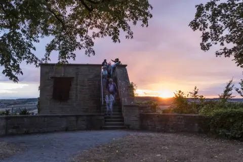 Menschen steigen die Treppen zum schlosskeller herrenberg hinauf, während die Sonne bei einem dramatischen Abendhimmel untergeht.