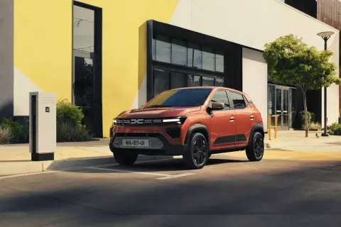 En orange Dacia står parkerad vid en laddstation framför en modern byggnad. Är Dacia bra eller dålig? Denna ser lovande ut!