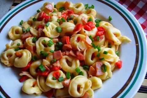 Pyszne tortellini co to jest? Makaron z kawałkami szynki, papryki i szczypiorku na talerzu.