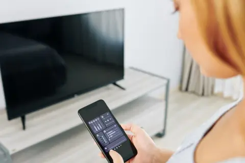 Kobieta trzyma smartfon z aplikacją do sterowania TV, obok stoi włączony telewizor.