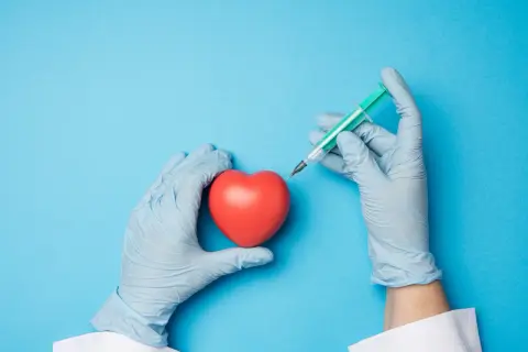 Mains gantées tenant un cœur rouge et une seringue, évoquant une intervention comme une coronarographie.
