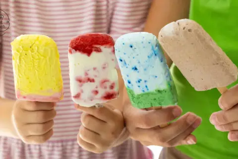 Vier Kinderhände halten bunte Fruchteis am Stiel: gelb, rot-weiß, blau-grün und braun.