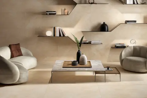 Ein modernes Wohnzimmer in warmen Beigetönen. Ein geschwungenes Sofa und ein Sessel laden zum Entspannen ein. Regale mit Büchern und Deko schmücken die Wand.