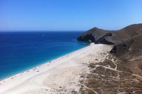 Playa de arena blanca y mar azul intenso, con acantilados rocosos y vegetación árida. Un velero navega en la distancia, un capricho del paisaje.