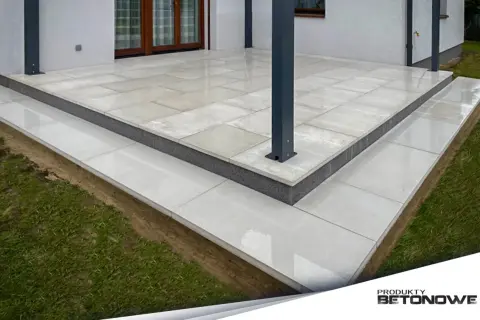 Nowoczesny taras z płyt betonowych, z podwyższonym stopniem i schodkami, otoczony zieloną trawą.