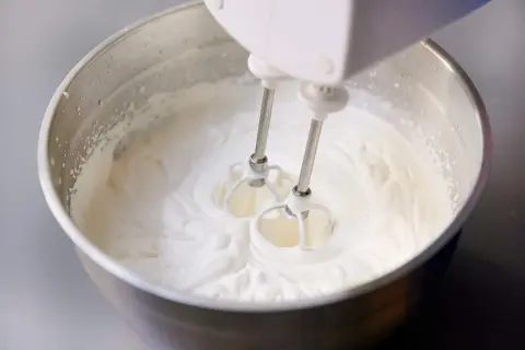 Schlagobers wird mit einem Handmixer in einer Edelstahlschüssel aufgeschlagen. Die cremige Masse ist bereit für süße Köstlichkeiten.