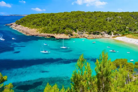 Bahía de aguas turquesas y cristalinas, con veleros y lanchas navegando. Al fondo, una playa de arena blanca y una frondosa vegetación que llega hasta la cala pudent.