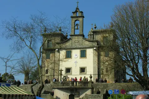 Edificio religioso con campanario y cruz. Gente en el balcón y en la plaza. Un árbol desnudo en primer plano.