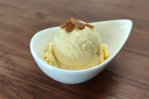Cremiges Eis mit Eigelb, garniert mit karamellisierten Stückchen, in einer weißen Schale auf Holzhintergrund.
