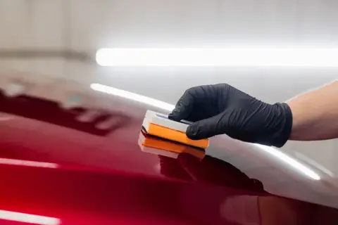 Ręka w czarnej rękawiczce nakłada preparat na błyszczący, czerwony lakier samochodu. Efekt jest olśniewający, niczym **kolor perłowy**.
