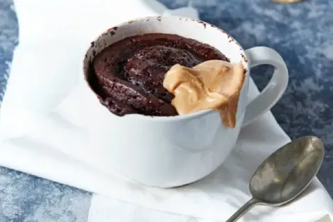 Schokoladiger high protein tassenkuchen mit Erdnussbutter-Topping in einer weißen Tasse, daneben ein Löffel.
