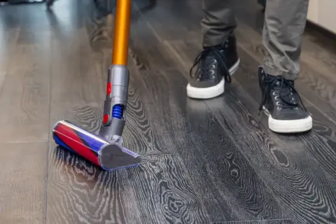 Persona aspira el suelo de vinilo con una aspiradora de mano. El suelo tiene un patrón de madera oscura.