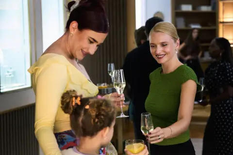 Två kvinnor och ett barn minglar med glas. Kvinnan i gult ler mot barnet, medan kvinnan i grönt håller upp sitt glas. Kanske en scen ur danska serier.