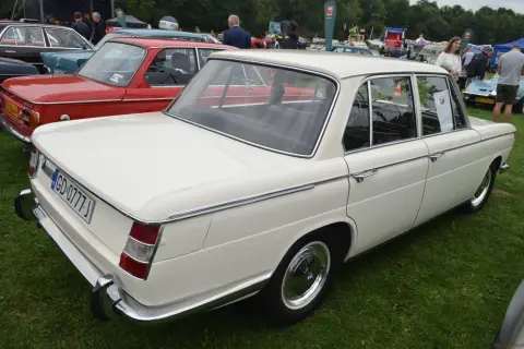 Białe BMW E10 z lat 70. prezentuje się elegancko na zielonej trawie, obok czerwonego klasyka.