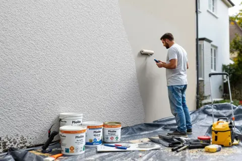Un homme peint un mur en pierre texturé en blanc avec un rouleau. Des pots de peinture sont au sol.