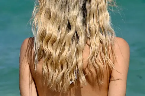 Blondes Haar mit Beach Waves im Wind, Schultern und Nacken einer Frau vor türkisblauem Wasser.