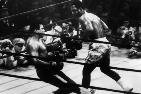 Joe Frazier fights in the ring, delivering a powerful blow as his opponent stumbles. The crowd watches intently.