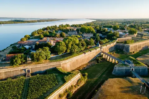 Vista aérea de la ciudadela de Blaye, con sus imponentes murallas de piedra, un puente arqueado y viñedos verdes junto a un río sereno.